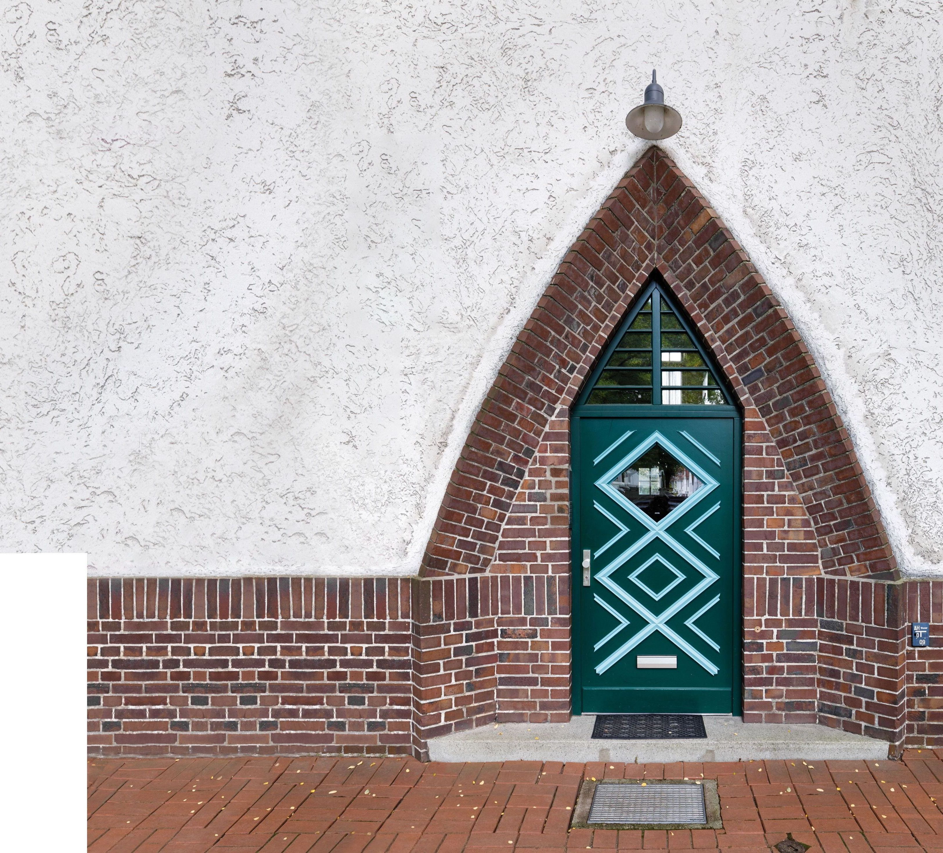Blick auf eine spitz zulaufende Rundbogen-Haustür in einer die Wand von unten halb mit Backstein eingefassten Fassade
