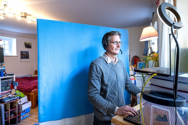 Ein Mann im provisorisch aufgebauten Homeoffice vor einer blauen Stellwand, die einen Einblick in das restliche Zimmer verhindert.