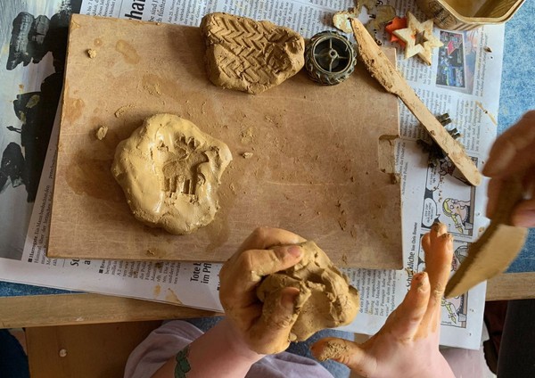 Kinderhände, die einen Klumpen Ton formen, und kleine Tonobjekte auf einem Holzbrett.