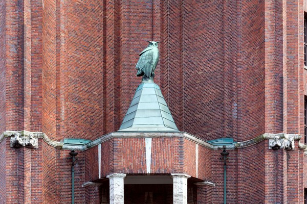 Eule aus angelaufenem Kupfer sitzt auf einem Dach eines runden Vorbaus, der einer ihn halbrund umschließenden Backsteinfassade vorsteht.