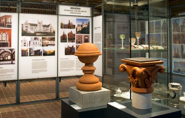 Blick in die Ausstellung Backsteinhistorismus mit Fassaden-Elementen von Backsteinbauten in Vitrinen und Fotografien von Gebäuden auf Tafeln.