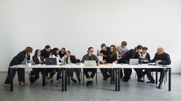 Eine Gruppe von Menschen sitzt und steht an einem langen Tisch, auf dem Laptops und Unterlagen verteilt sind
