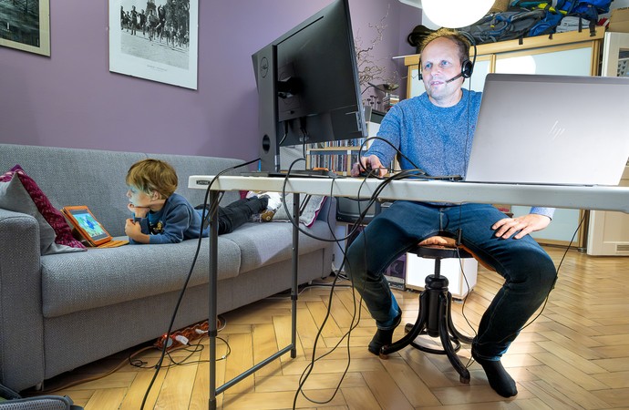 Ein Mann arbeitet an einem Computer an seinem Schreibtisch; auf dem Sofa daneben liegt ein Kind und spielt an einem Tablet.